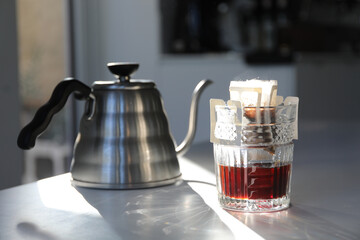 Glass with drip coffee bag and kettle on light grey table, closeup