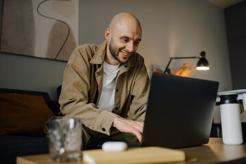 A smiling man is working on a laptop in his apartment