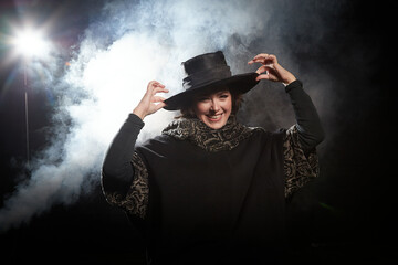 Portrait of a woman in black warm coat and black hat on dark background with smoke. Female model girl posing outdoors in spring, autumn or winter day