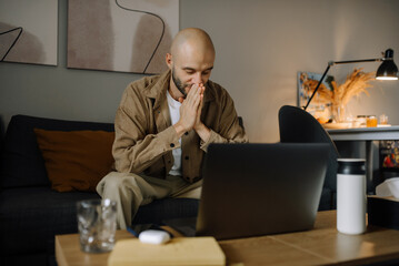A man is tired of working on a laptop at home. Stressed depressed freelancer touching his head, feeling pain in eyes.