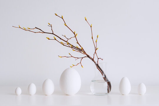 Minimalistic Easter still life. A glass vase with a branch on which colorful Easter eggs hang on a white background. Interior photography.