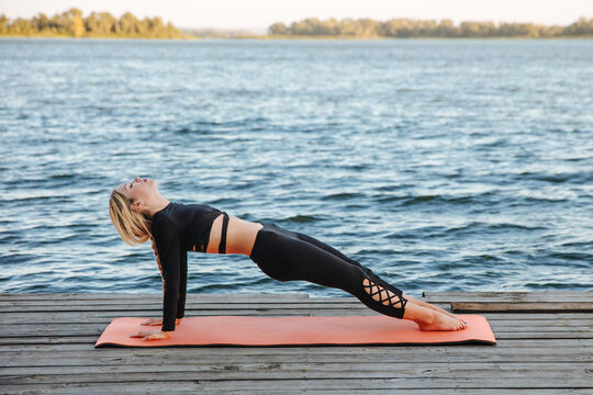 A young woman is doing a reverse plank on a yoga mat by the river.