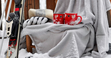 two red mugs with heart shaped on a plaid put on a chair  in  snow andski equipment on terrace