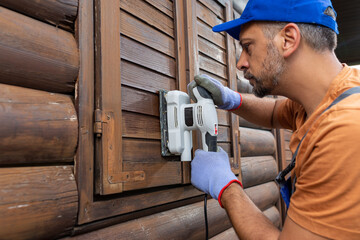 Worker sanding a wooden window shutters on house exterior.