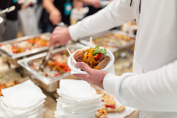 Close up of man holding plastic plate and scooping food at buffet table catering in hotel or...