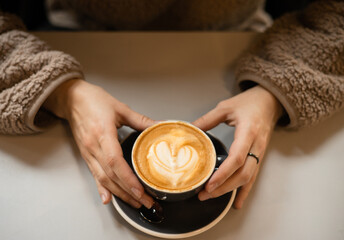 Thin female fingers hold a cup of coffee with heart shaped latte art foam. Close up cup of coffee with cream in coffee shop.