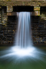 Small artificial cascade in Iserlohn Sauerland Germany near “Rupenteich“, a pond in an old park and recreation area with natural creeks and historic brick walls. Longtime exposure waterfall.