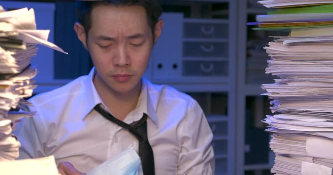 Sick asian business man is sitting at desk covered with stack of paperwork and wearing face mask. Unwell alone male is coughing and feeling exhausted while working hard at late night. Close up shot.