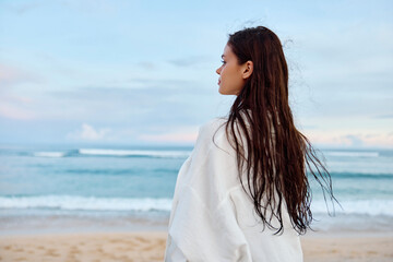 Fototapeta na wymiar Happy tanned slender woman in white swimsuit shirt and denim shorts walks on the beach with her back to the camera on the sand by the ocean with wet hair after swimming, sunset light