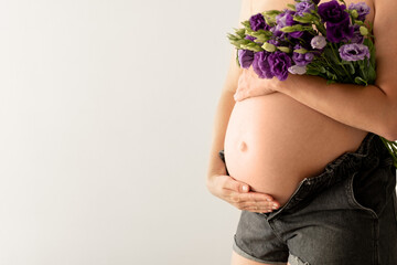 Cropped shot of unrecognizable pregnant woman,perfect skin,hiding breast with flower bouquet touching,caressing tummy,belly on white background.Beautiful pregnancy in spring,breastfeeding.Copyspace.