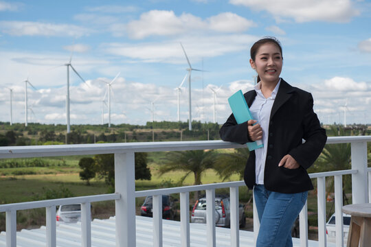 person holding folder. Asian woman in white helmet working with digital tablet at renewable energy farm. Female inspector controlling functioning of wind turbines outdoors.
