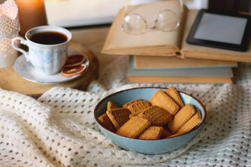 Vintage books, reading glasses, e-reader, cup of tea, bowl of biscuits and lit candle on the table. Selective focus.