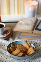 Vintage books, reading glasses, e-reader, cup of tea, bowl of biscuits and lit candle on the table. Selective focus.