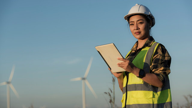engineer in helmet with a clipboard on wind turbine on background. Asian woman in white helmet working with digital tablet at renewable energy farm. Female inspector controlling functioning of wind tu