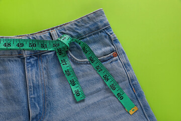 Jeans and measuring tape on light green background, top view. Weight loss concept