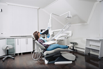 Young woman patient in checkered shirt with perfect straight white teeth waiting for dentist in dental chair and smiling relaxed, ready for a check-up. Beautiful woman smile