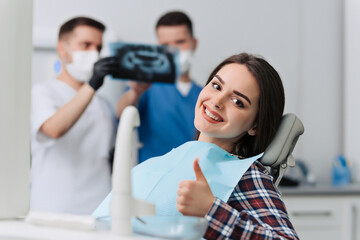 Portrait of handsome female patient showing thumb up with dentists analysing x-ray on background.