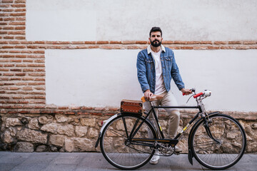 Obraz premium Young man in denim jacket holding his vintage classic bicycle while looking his mobile phone in front a brick wall.