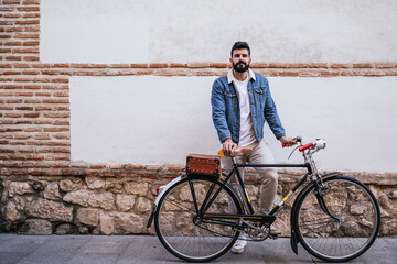 Young man in denim jacket holding his vintage classic bicycle while looking his mobile phone in...