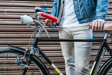 Obraz premium Cropped image of a young man in denim jacket holding his vintage classic bicycle in front of the closing shutter of a shop.