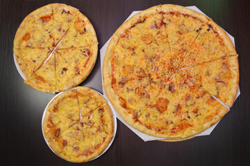 Three delicious cheese pizzas with ham, sausage, tomatoes on black table, three different sizes for...