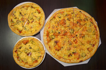 Three delicious fresh cheese pizzas with mushrooms, pickles, sausage on black table, different sizes for serving