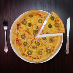 Delicious fresh cheese pizza with ham, olives on black table, surface, on white plate, fork, knife nearby, top view