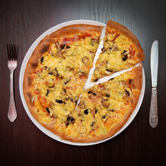 Delicious fresh cheese pizza with mushrooms, ham on black table, surface, on white plate, fork, knife nearby, top view