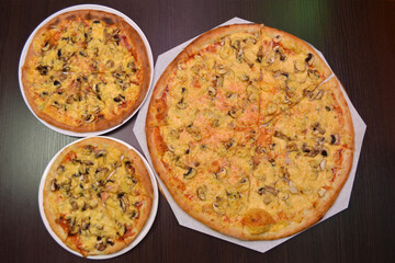 Three delicious fresh round cheese pizzas with mushrooms, ham on a black table, surface, on a white plates, three different sizes for serving in a pizzeria