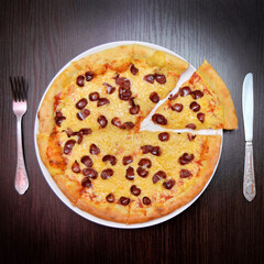 Delicious fresh round cheese pizza with smoked sausages on black table, surface, on white plate, fork and knife nearby, top view