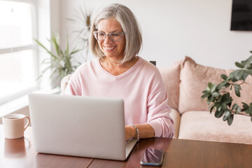 Happy middle age senior woman sitting at the table at home working using computer laptop