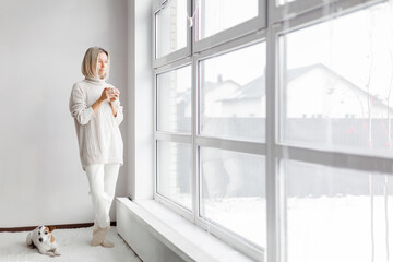 Adult woman stands in white room next to a panoramic window and drinks a cup of coffee in the morning