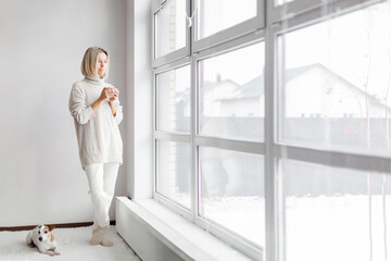 Adult woman stands in white room next to a panoramic window and drinks a cup of coffee in the...
