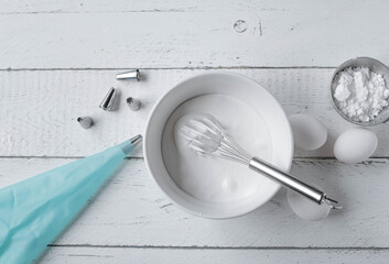 Royal icing or egg white glaze for decorating a cake or cookies. Served with whisk in a bowl and kitchen utensils such as piping bag  on white background table. Top view