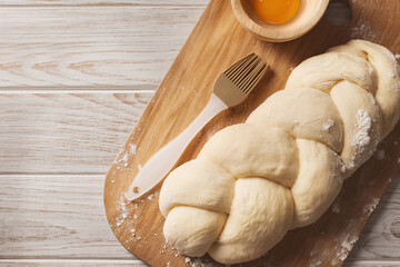 Raw braided bread, egg yolk and pastry brush on white wooden table, top view with space for text. Traditional Shabbat challah