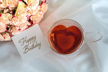 Herzförmige Teetasse aus Glas mit rotem Tee gefüllt auf weißem Satinstoff, hellrosafarbene Rosen oben links und ein weißer Notizzettel mit dem Schriftzug  Happy Birthday  im Bild, Topview, horizontal 