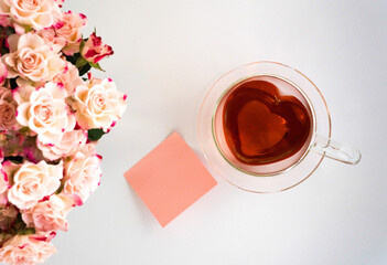 Herzförmige Teetasse aus Glas mit rotem Tee gefüllt auf weißem Hintergrund hellrosafarbene Rosen links und ein rosafarbener Notizzettel blanko im Bild, Topview, horizontal,