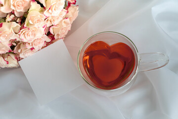 Herzförmige Teetasse aus Glas mit rotem Tee gefüllt auf weißem Satinstoff, hellrosafarbene Rosen...