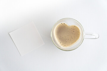 Herzförmige Tasse aus Glas mit Kaffee gefüllt vor weißem Hintergrund, weißer Notizzettel blanko...
