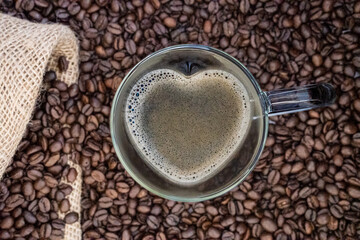 Herzförmige Tasse aus Glas mit Kaffee gefüllt,im Hintergrund unscharf Kaffeebohnen und teilweise ein Jutesack zu sehen , Topview, horizontal