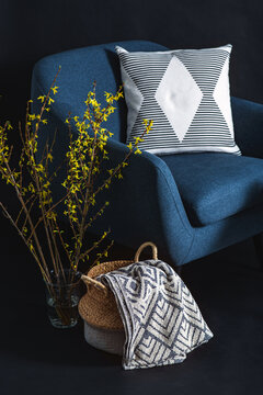interior and home decor concept - close up of blue chair with pillow, blanket in wicker basket and tree branches in vase over black wall