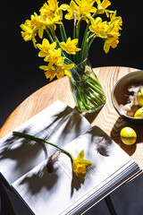 interior, holidays and home decor concept - close up of easter eggs in bowl, daffodil flowers and magazine on table in dark room