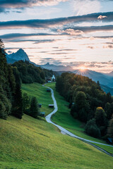 Sonnenuntergang in den Alpen in Bayern mit Straße, grüner Wiese und Kapelle.