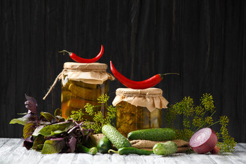 Pickled cucumbers in glass jars and spices and vegetables for preparation of pickles on old wooden background.