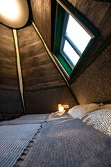 Double bed in bedroom of pyramid shape wooden cabin. Pyramid shape walls of bungalow from inside. Small room, angled walls, window light, ceiling lamp, tree trunks corners, made bed.