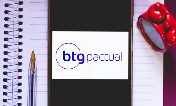 A logo do banco BTG Pactual na tela de um telefone celular. Um caderno na composi&ccedil;&atilde;o.