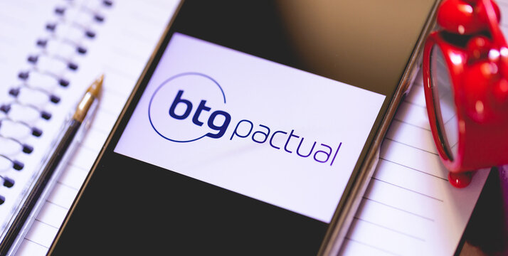 A logo do banco BTG Pactual na tela de um telefone celular. Um caderno na composi&ccedil;&atilde;o.