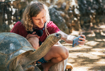 Smiling tourist boy making a selfie using cell phone with Aldabra giant tortoise endemic species - one of the largest tortoises in the world in the zoo nature park on Mauritius island. - obrazy, fototapety, plakaty