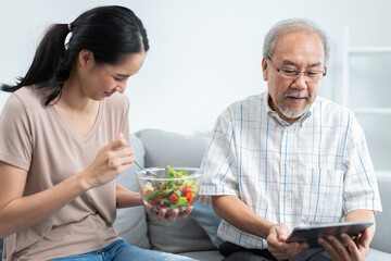 Young Asian daughter serving and feeding food to senior father while he using and playing on digital tablet in the living room. family love and support to elderly people. 