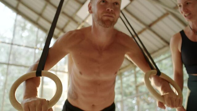 Bare-chested young man doing push-ups using suspension ropes during a gym session with an instructor